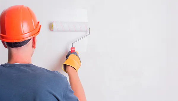 Profissional de reforma usando capacete de segurança laranja e camisa azul pintando parede branca com rolo, representando serviços de manutenção, construção civil e renovação de ambientes.