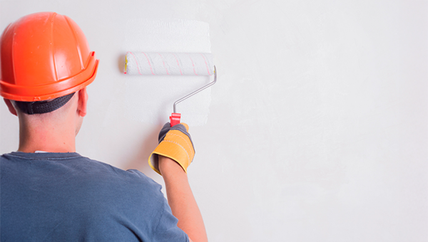 Profissional de reforma usando capacete de segurança laranja e camisa azul pintando parede branca com rolo, representando serviços de manutenção, construção civil e renovação de ambientes.
