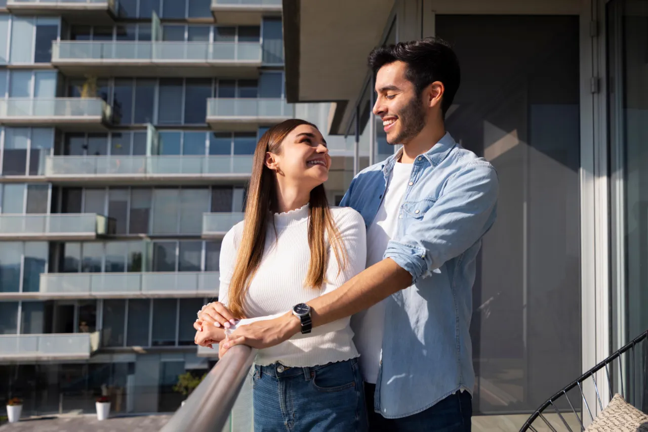 Imagem de um jovem casal feliz na varanda de seu novo lar, após a aquisição em um consórcio de apartamento.