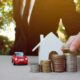 Homem empilhando moedas ao lado de miniatura de casa e carro, ilustrando o investimento em consórcio