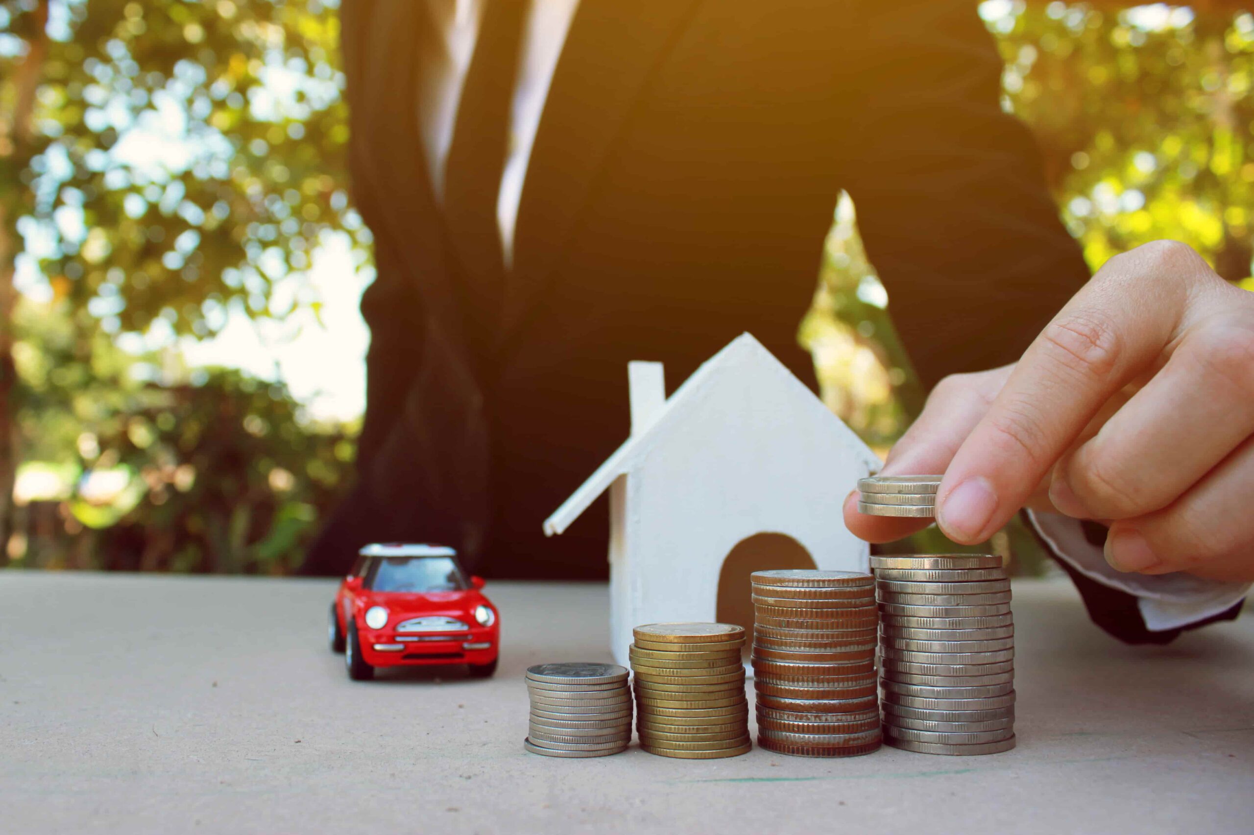 Homem empilhando moedas ao lado de miniatura de casa e carro, ilustrando o investimento em consórcio